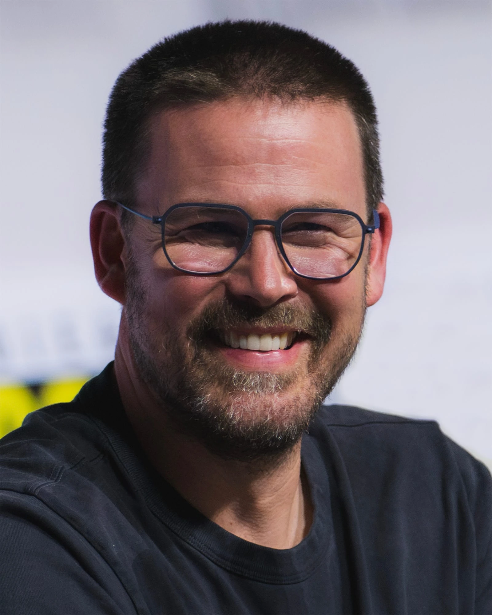 Zach Cregger sonríe a cámara, con gafas de montura metálica, barba corta y camiseta oscura