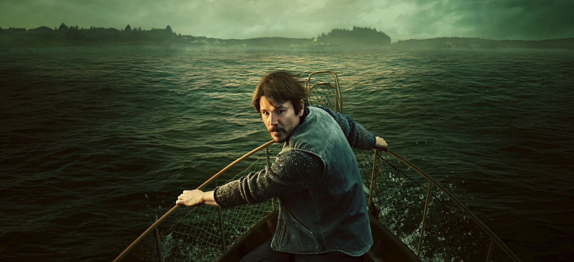 Josh Hartnett, con chaleco y jersey de lana, se agarra a la barandilla de proa de una barca y mira hacia atrás sobre un mar verdoso, con la costa en penumbra al fondo