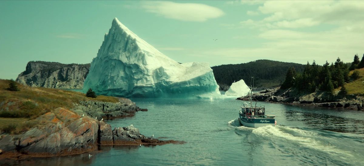 Un barco de pesca avanza por una ensenada rocosa hacia un gran iceberg, bajo un cielo claro