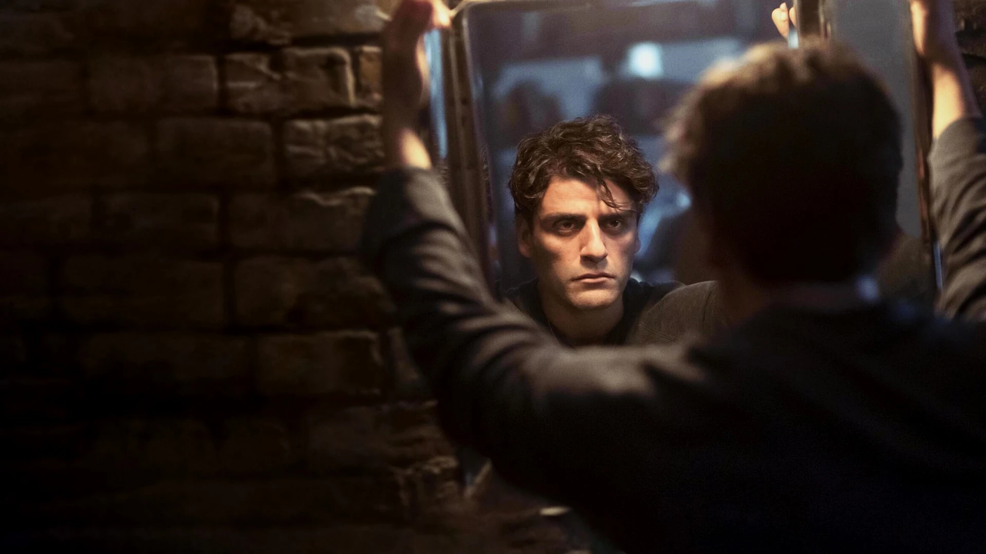 Oscar Isaac, serio, mira de frente desde el otro lado de un marco, mientras una figura de espaldas se apoya en él en una habitación en penumbra con pared de ladrillo