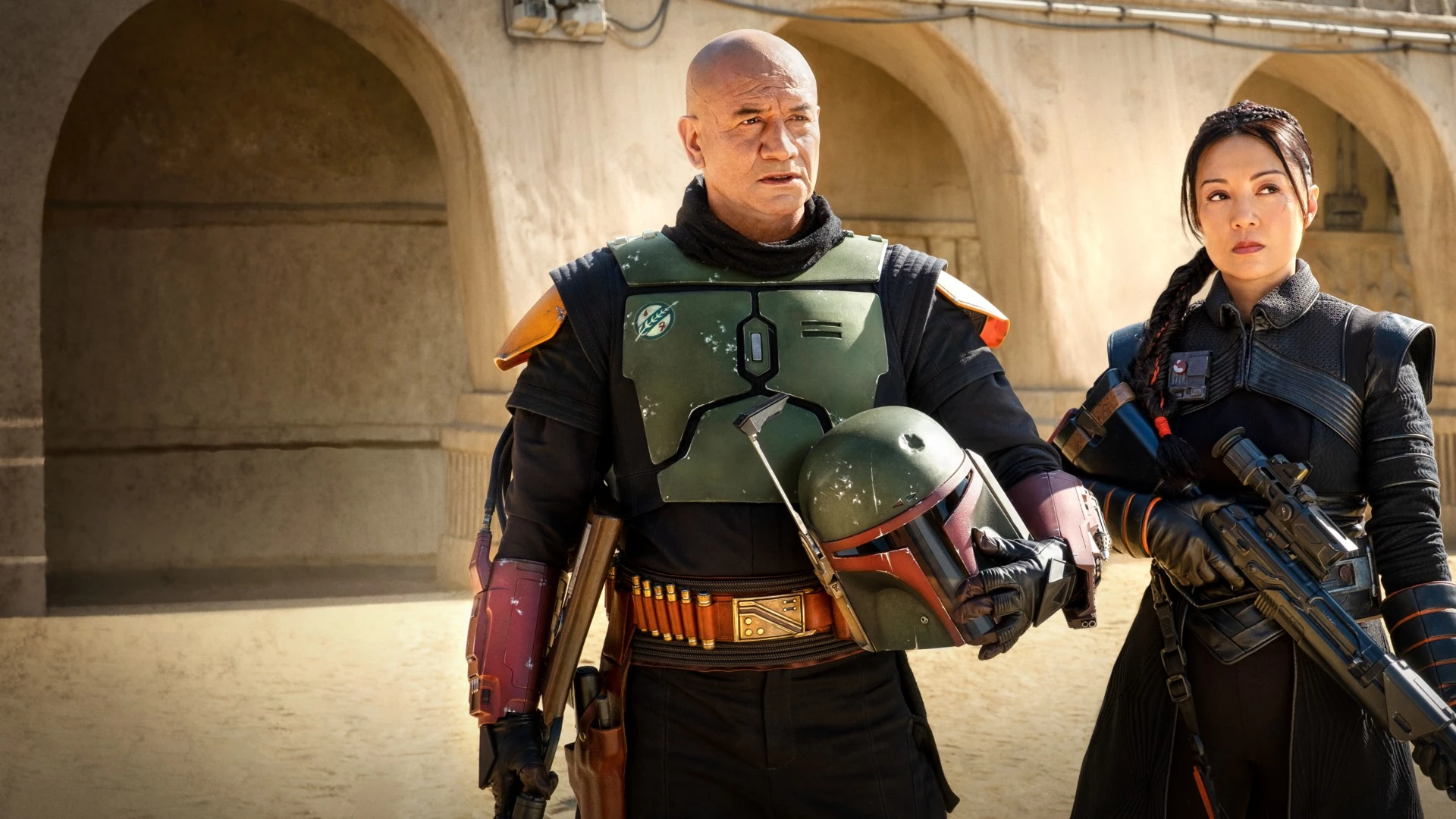 Boba Fett, con armadura verde y el casco bajo el brazo, y Fennec Shand, con un rifle, caminan bajo unos arcos de piedra de color arena