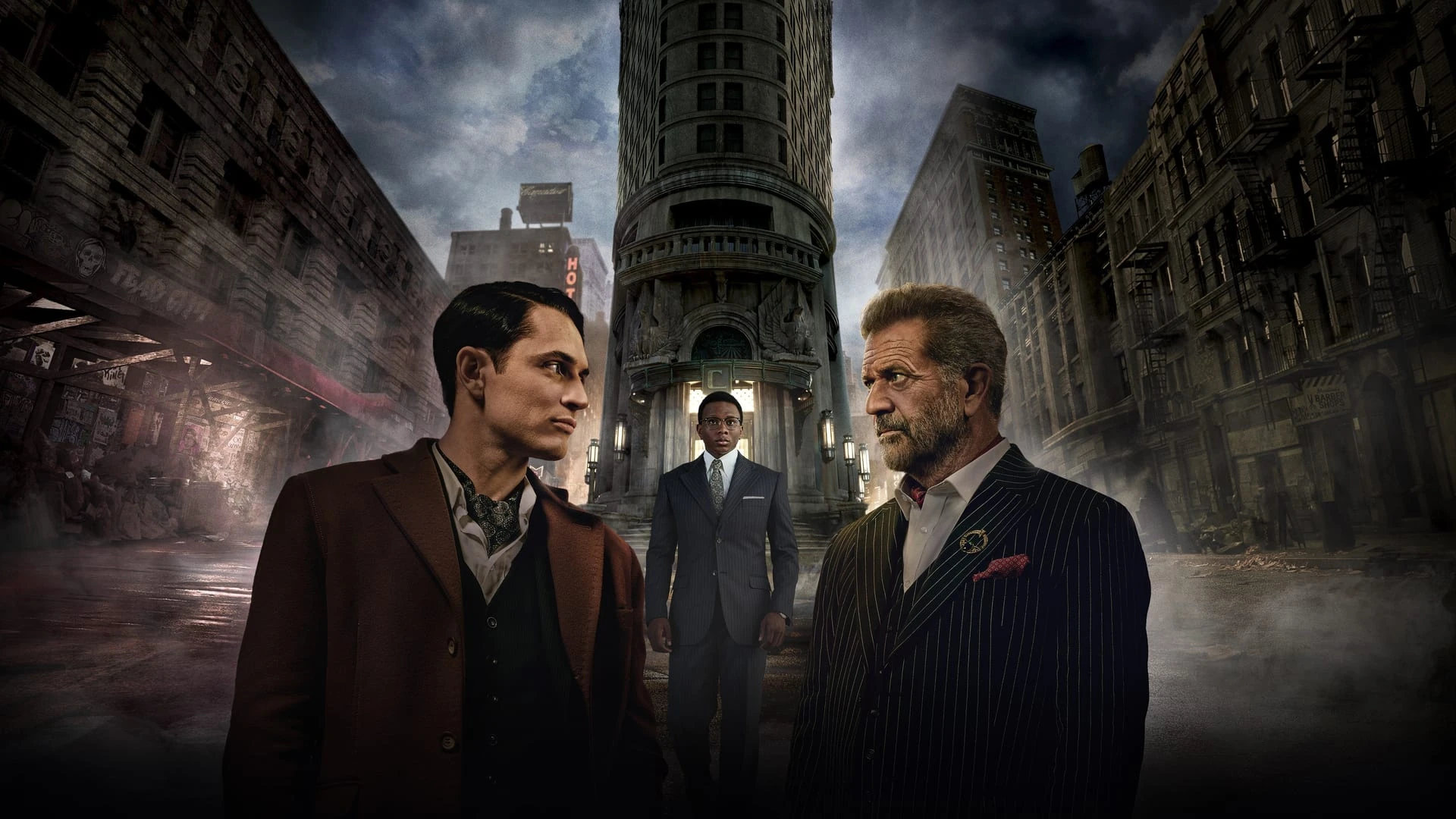 Colin Woodell y Mel Gibson, de traje, se miran frente a frente en una calle neoyorquina con humo; entre ellos, al fondo, un joven de traje gris ante la entrada de un edificio alto y estrecho
